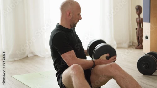 4K video captures man dressed black doing dumbbell russian twist exercise at home living room. Footage illustrates strength training, sport, healthy lifestyle concept.