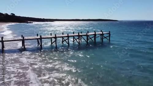 A wooden pier extends into the sea, framed by a beach and a distant shoreline under a clear sky