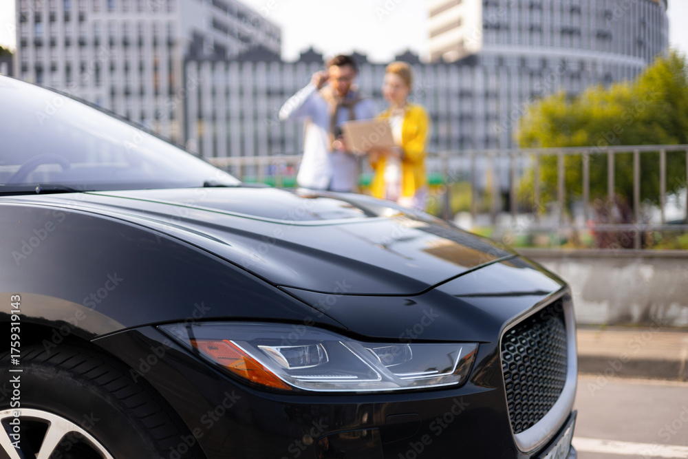 Fototapeta premium A sleek black car is the focus, with a blurred couple in the background near a city building.