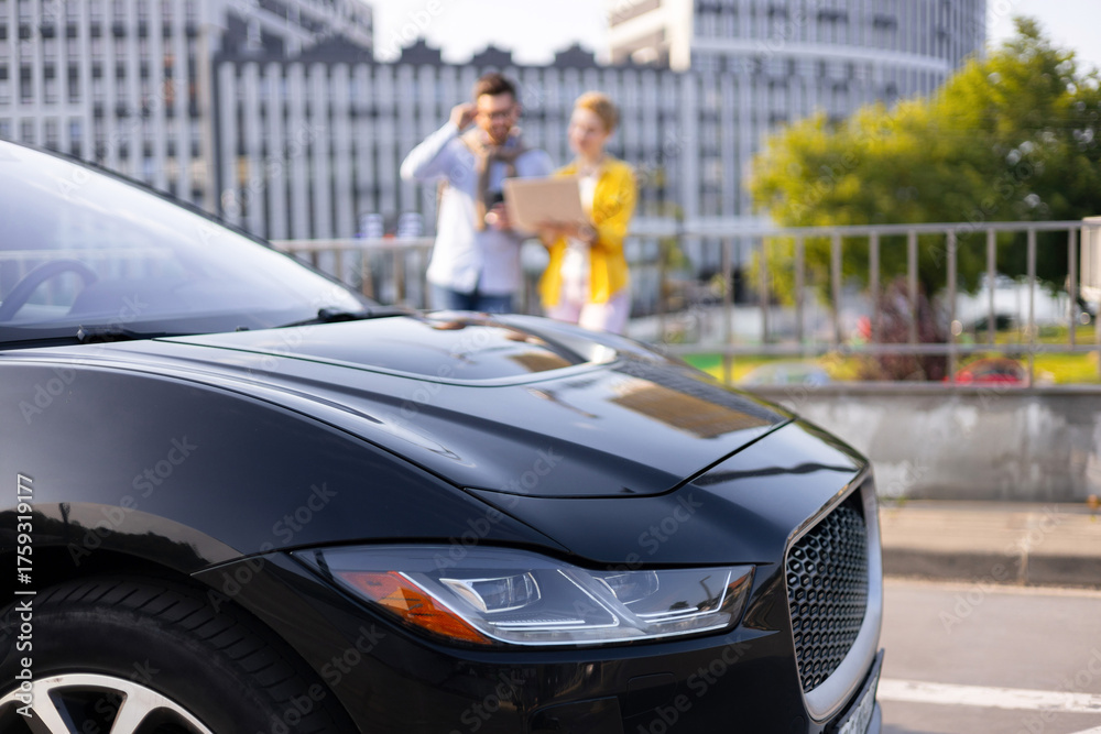 Fototapeta premium A sleek black car is the focal point, with a blurred background featuring two people and a cityscape.
