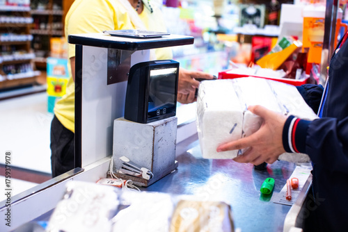 Selective focus to salesperson holds a product in his hand to scan a QR code to pay at the counter in a supermarket. Cashier at supermarket checkout counter. A barcode scanner sits on the counter.