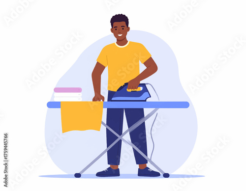 A smiling Black man in a yellow t-shirt and blue pants irons clothes on an ironing board, with a stack of folded laundry beside him.