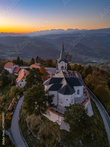 Aerial view of the Pilgrimage Church of the Birth of Mary and its defensive wall atop the 852m high Holy Mountain of Zasavje at sunset, surrounded by autumnal forests.