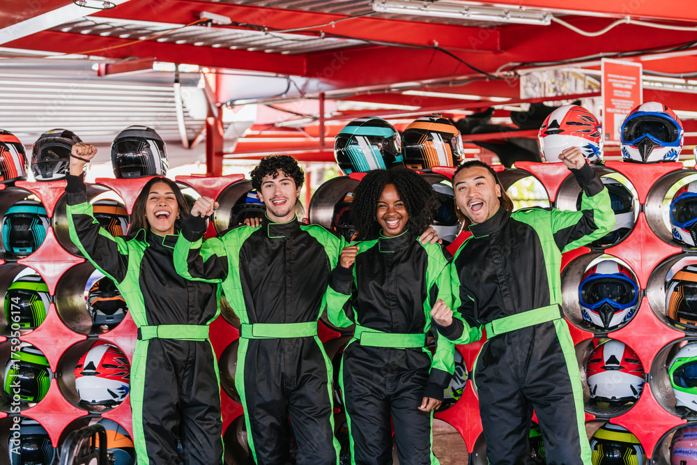 Obraz premium Group of excited friends in racing suits raising fists, celebrating a go kart victory or successful race at an indoor track