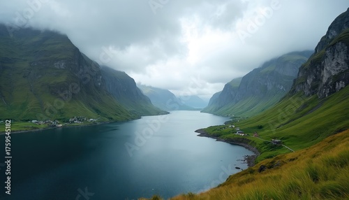Panoramic vista of Norwegian fjord landscape under overcast skies. Rich green hills descend to calm blue waterway. Small settlements dot shoreline within expansive wild region. Presents serene
