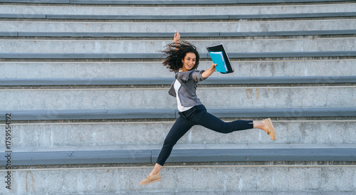 Enthusiastically leaping on outdoor steps with a folder in hand, a woman exudes joy and carefree spirit, embracing life’s spontaneity.