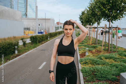Canvas Print A woman in sporty attire walks confidently down a tree-lined street, brushing back her hair, exuding casual poise and energy in a vibrant urban setting