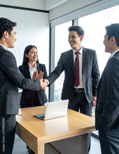Group of professionals in formal attire shake hands around a table with a laptop, an office setting near windows