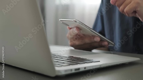 Man using smartphone and laptop at desk, close up of hand multitasking with technology in modern workspace, typing for communication and digital lifestyle