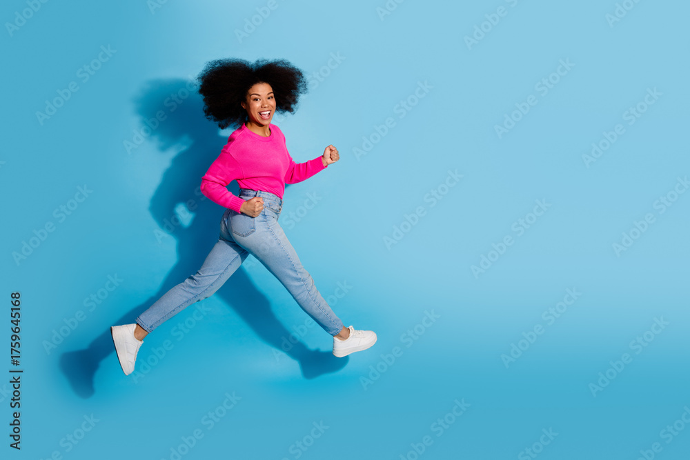 Obraz premium Cheerful young woman jumping energetically in pink sweater and blue jeans against a vibrant blue background showcasing joy