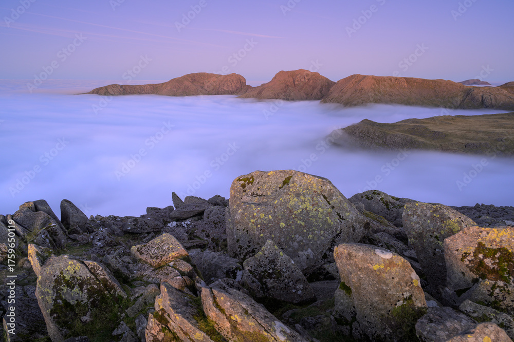 Fototapeta premium Bowfell summit rocks with incredible view looking over to Scafell Massif mountain range with a thick cloud inversion below. Lake District, UK.