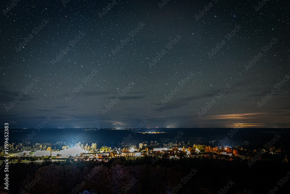 Fototapeta premium A clear night sky filled with countless stars above a distant landscape with faint city lights and clouds illuminated by a warm horizon glow.