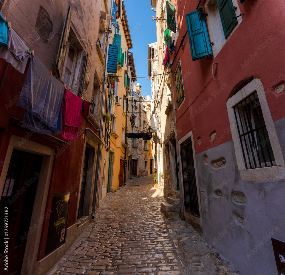 Obraz premium Rovinj, Croatia - August 15, 2025: Narrow Alley in Rovinj – colorful old buildings with open shutters and laundry hanging across a sunlit cobblestone street in the charming old town of Rovinj, Croatia
