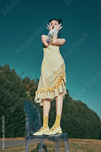 Young woman in yellow dress standing playfully on transparent chair outdoors.
