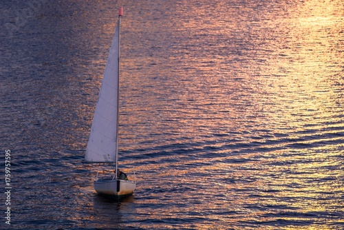 Bilde på lerret Sailing into the Sunset: A lone sailboat glides effortlessly across the shimmering water under a sky painted with the golden hues of the setting sun, evoking a sense of serene freedom and adventure