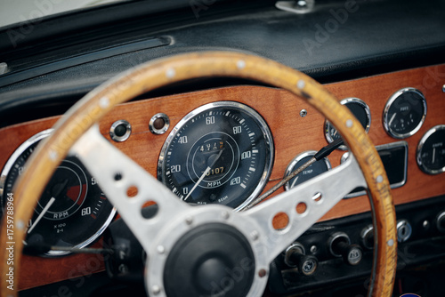 Close-up on the interior of a classic open-top car, featuring a wood-rimmed wheel, speedometer, and other gauges on a wooden dashboard.