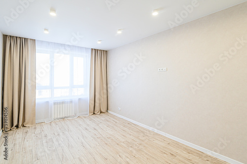 Bright, empty room with wood floors, beige walls, and a large window covered by light curtains. Ceiling spotlights illuminate the space