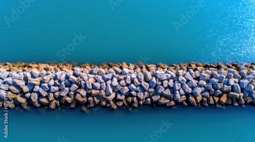 Aerial view of a rocky breakwater extending into a serene turquoise sea, with gentle waves lapping