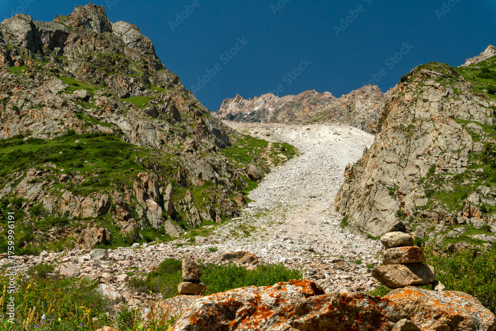 Naklejka premium Rocky mountain pass with steep slopes, scattered cairns, scree field, lush green patches, blue sky, rugged terrain, alpine environment, popular hiking and mountaineering destination, summer season