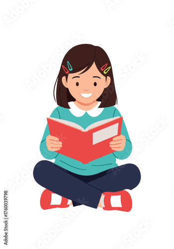 A young girl happily reads a red book while sitting cross legged.