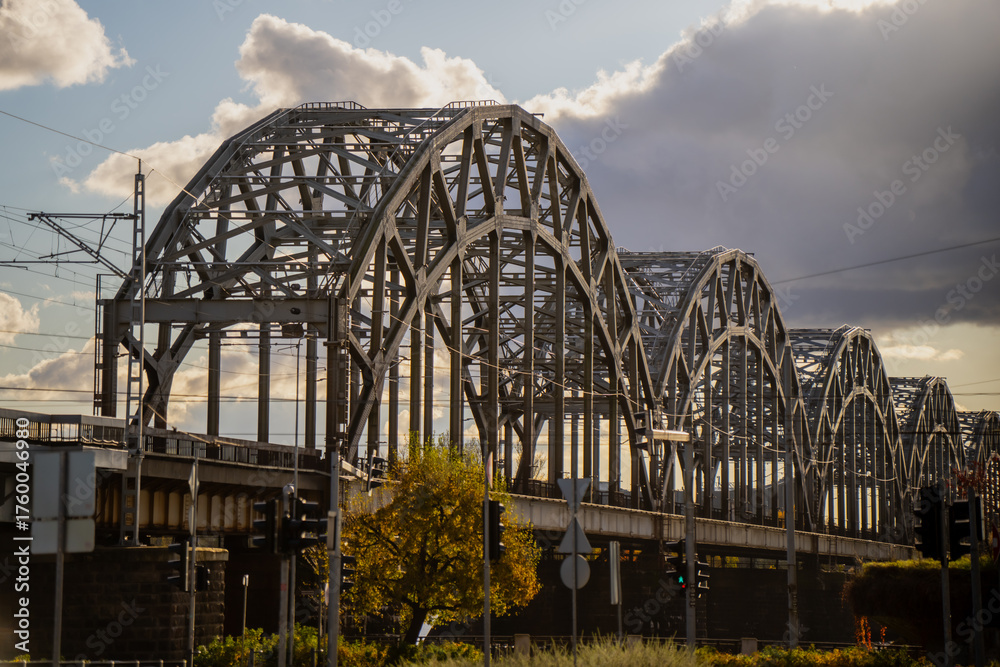 Obraz premium Riga steel railway bridge with repeating arches under dramatic cloudy sky representing industrial engineering, architectural design, and urban transportation infrastructure
