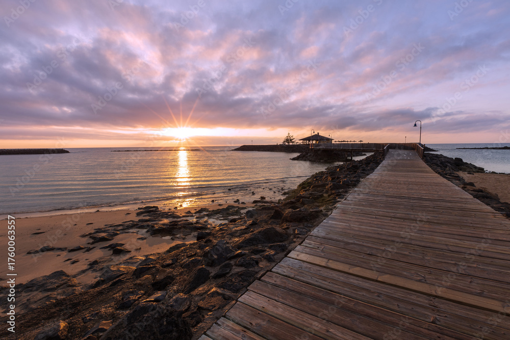Obraz premium Beach in Fuerteventura, Coralejo. In the foreground, rocks on the beach and in the background, a small bar on the pier at sunrise
