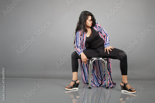 A female dancer sits on a metal stool, striking a lively, expressive pose as her striped long coat swirls around her. Perfect for fashion, lifestyle, empowerment, and creative movement concepts.