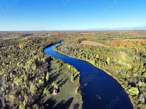An aerial drone view captures the Indian River in Ontario as it meanders through an expansive forest landscape exhibiting vibrant autumn foliage under a clear blue sky