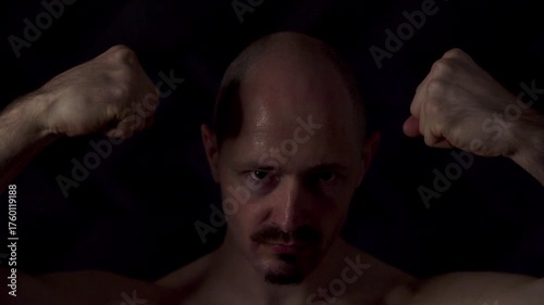 Close-up portrait of an athletic man against a dark background. He raised his arms and tensed all his muscles, showing his strength and power. 
