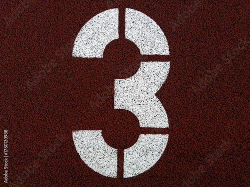 White number three painted on red running track surface, close-up of athletics field marking with textured background representing ranking, third place, progress, and sports competition