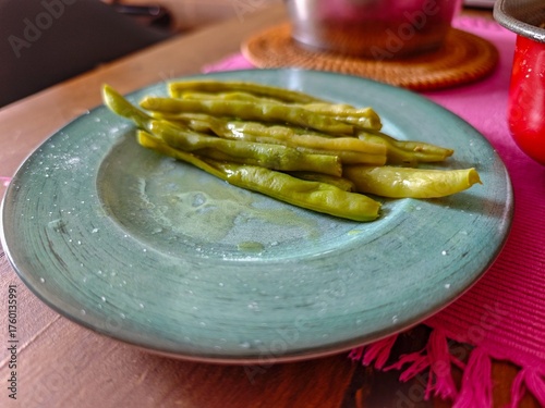A beautiful presentation of cooked green beans arranged artistically on a bluish-green ceramic plate. The homey scene is complemented by a pink cloth and a straw holder, conveying warmth and comfort. 