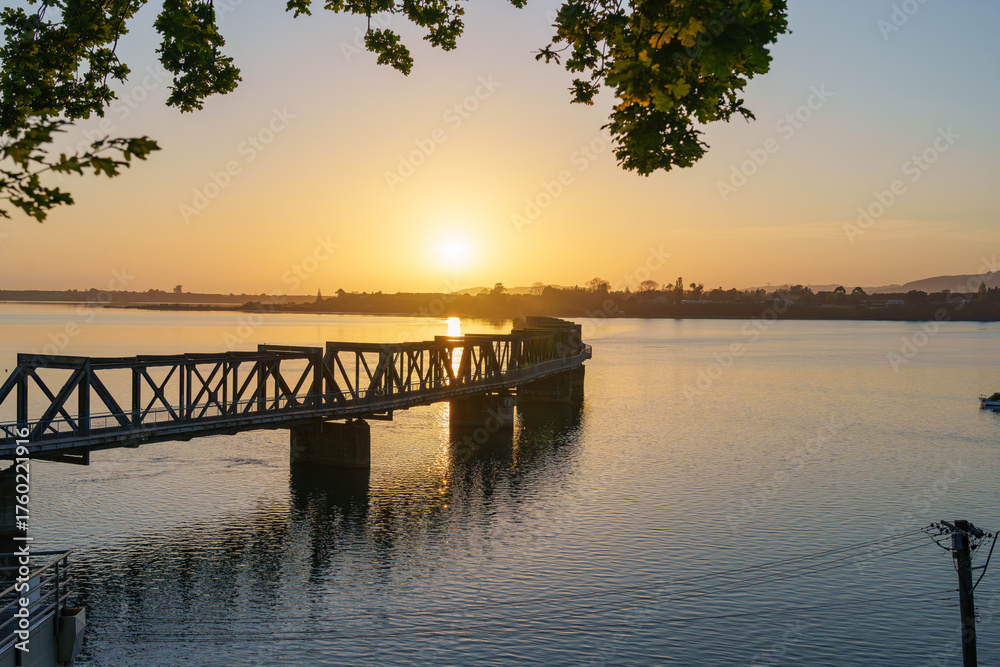Fototapeta premium Sun rises over Tauranga harbour and historic railway bridge on calm morning