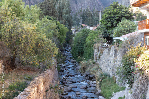 The Valley's Lifeline; a photograph of a mountain stream flowing through a narrow valley village, framed by steep rocky cliffs and lush vegetation.
