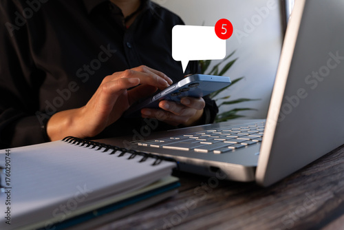 A woman sitting at a desk with a laptop using her cell phone. She's checking her phone and sees a speech bubble with a notification that says 