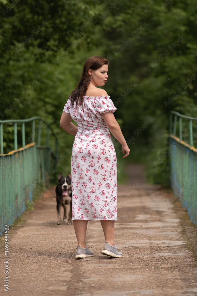 Naklejka premium Young woman with long brown hair wearing a floral dress stands beside a tree, gazing thoughtfully into the distance, surrounded by lush greenery and natural beauty