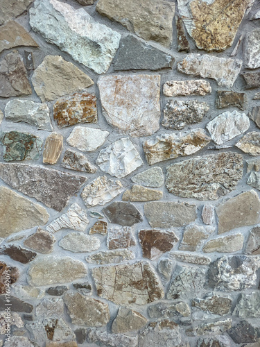 A detailed, full-frame shot showcasing the rugged, irregular texture of a stone wall constructed with various sizes of natural, multi-toned fieldstones and visible mortar joints.