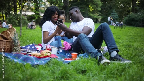 Cute couple in love and little daughter sitting beside each other on picnic blanket. Young African man holding black smartphone in hands, showing photos, smiling.