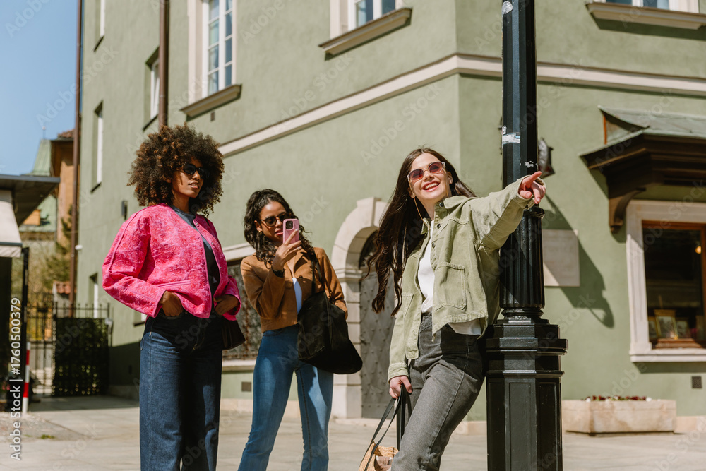 Obraz premium A woman leans on a street lamp and points to the side while laughing and standing next to her are two female friends, one of whom is holding a phone