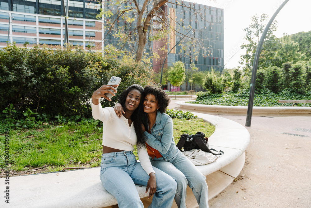 Naklejka premium A female student hugs a female student sitting next to her while they smile and pose in front of a phone that one of them is holding