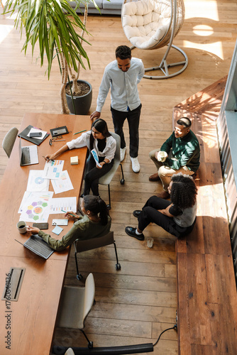 A male worker stands next to a group of four workers sitting at a table and on a bench