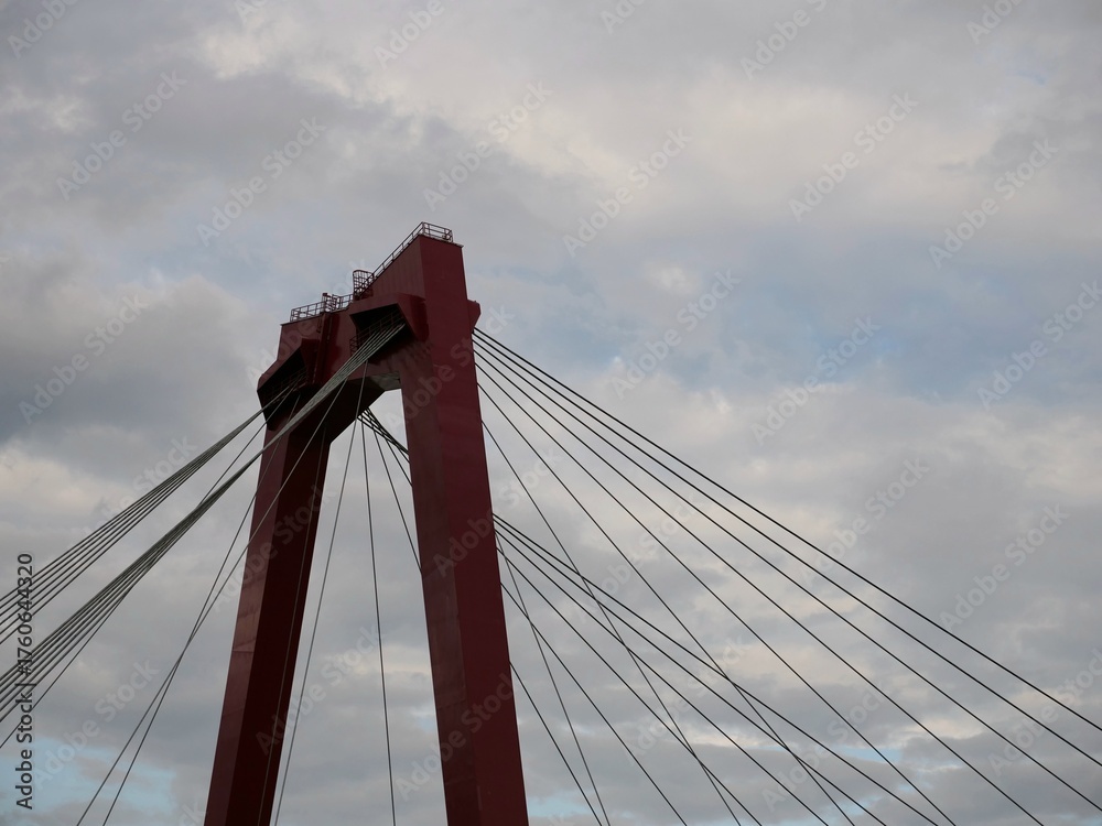 Fototapeta premium Red suspension bridge tower against dramatic cloudy sky, architectural close-up with steel cables. Engineering marvel, iconic structure, urban infrastructure, minimal color contrast.