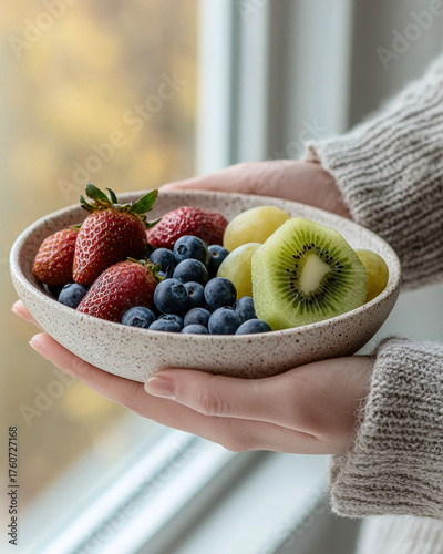 Fresh and vibrant fruit bowl featuring strawberries, blueberries, green grapes, and kiwi, held gently in hands. natural light enhances colors, creating warm and inviting atmosphere