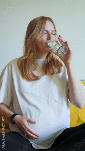 Video of pregnant woman sitting on bed, holding her belly and drinking water. Cozy home atmosphere with soft light. Concept of healthy lifestyle during pregnancy, hydration, self-care, and motherhood.