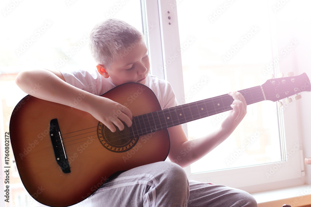 Fototapeta premium A blond boy plays the guitar on the windowsill by the window.