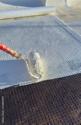 Worker applying white polyurethane waterproof coating on flat roof surface with paint roller.