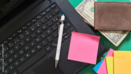 Desk essentials with laptop, colorful post-its, pen and leather wallet with dollars inside on green background
