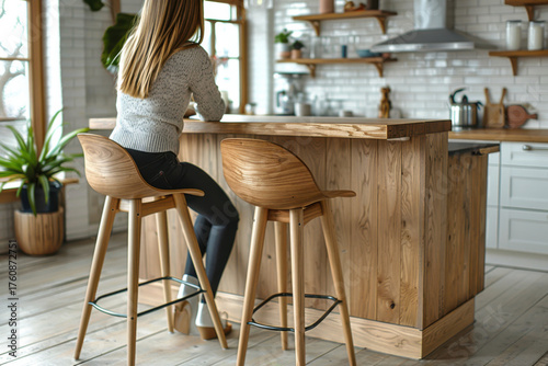 In a bright minimalist kitchen made of pale ash wood, a person sits on a stool at the countertop. Natural light streams through the windows, enhancing the warm ambiance of the space