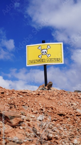 Danger mines sign standing in reddish, rocky terrain under a bright blue sky, symbolizing the lasting threat of explosive devices in post conflict regions and the need for demining efforts