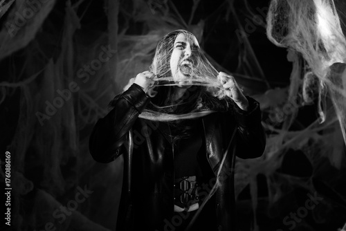 Sinister woman with Halloween vampire makeup trapped in spider web trying to scare. lady in black leather coat against black background