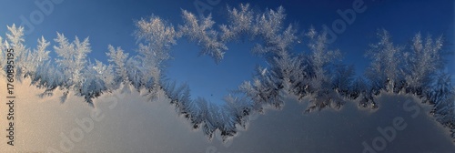 Delicate frost patterns adorn the edge of a windowpane, forming a heart shape against a serene blue sky during early morning light.